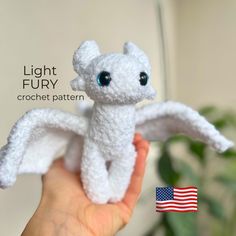 a hand holding a small white crochet dragon with blue eyes and wings, in front of an american flag