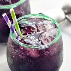 two glasses filled with purple liquid and ice on top of a white table next to spoons