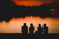 three people are sitting by the water at sunset