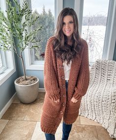 A person stands in a cozy room, wrapped in a long brown cardigan and jeans, with a crochet crop top pattern subtly displayed beside the knit blanket and potted plant.