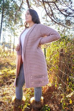 a woman standing in the woods with her hands on her hips, wearing boots and a pink cardigan