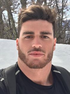 a man with a beard standing in front of some snow covered hills and trees, looking at the camera