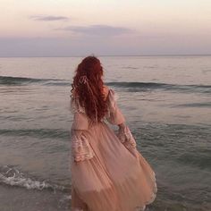 a woman walking into the ocean with her long red hair in a flowing pink dress