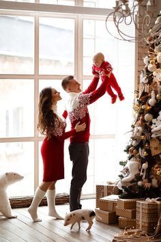 a man and woman standing next to a christmas tree holding a baby in their arms