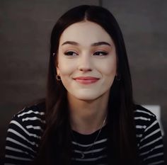 a woman with long black hair wearing a striped shirt and smiling at the camera while sitting in front of a gray wall