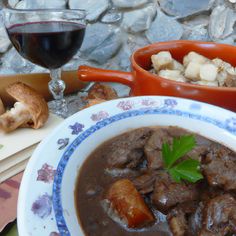 a bowl of beef stew next to a glass of red wine on a table with bread and croissants