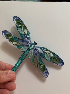 a hand holding a paper sculpture of a dragonfly on a white surface with green and blue swirls