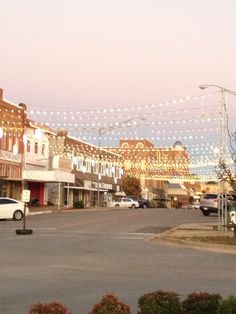 an empty street with cars parked on both sides and lights strung over the buildings in the background