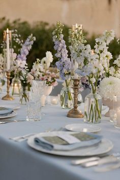 the table is set with white and blue flowers in vases, candles, and plates