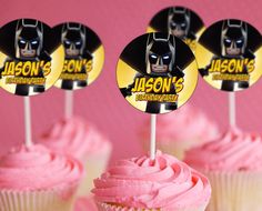 cupcakes with pink frosting and batman stickers on top are displayed in front of a pink background