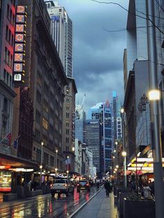 a city street with tall buildings and people walking on the sidewalk in the rain at night