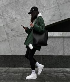 a woman walking down the street looking at her cell phone while wearing a green coat and white sneakers