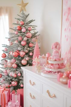 a decorated christmas tree with pink ornaments and presents in front of it, next to a white dresser