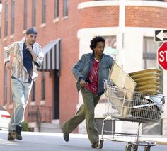 two men are walking down the street with a shopping cart full of boxes and papers