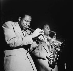 two men in suits are playing the trumpet while one man is looking at something behind them