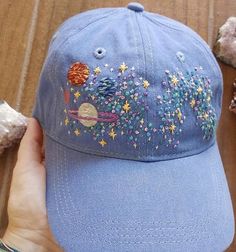 a hand holding a blue baseball cap with space and stars embroidered on it, next to shells