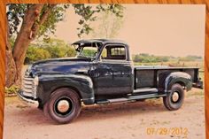 an old black truck parked next to a tree on a dirt road in front of a wooden frame