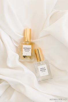 two bottles of perfume sitting next to each other on a white cloth covered bed sheet