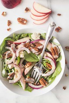 a white bowl filled with lettuce, apples and pecans next to a fork