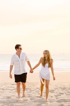 a man and woman holding hands while walking on the sand at the beach in front of the ocean