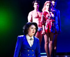 a woman in a blue jacket and skirt standing on stage with other people behind her