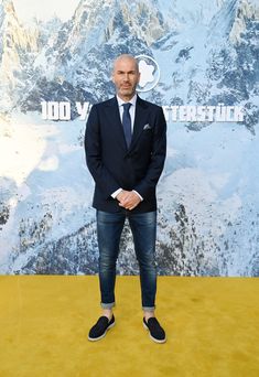 a man in a suit and tie standing on a yellow carpet with mountains behind him