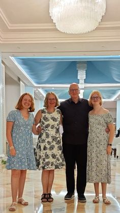 three women and one man are standing together in a hotel lobby with chandelier overhead