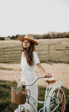 a woman wearing a hat standing next to a blue bicycle in a field with grass