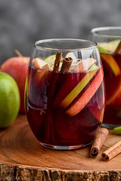 two glasses filled with apple cider sitting on top of a wooden table next to apples and cinnamon sticks