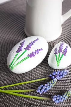 two rocks with lavender painted on them sitting next to a coffee mug and some flowers