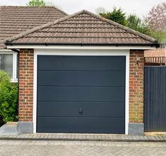 a grey garage door in front of a brick house with a black fence and bushes