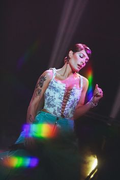 a woman with tattoos on her arm standing in front of colorful lights and holding a microphone