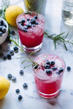 two glasses filled with blueberry lemonade and rosemary garnish on a marble table