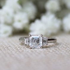 an engagement ring with three stones on it and white flowers in the backgroud