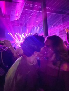 two people kissing each other in front of purple and green lights at a concert or party