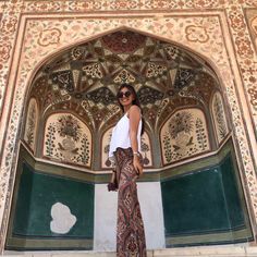 a woman standing in front of a wall with ornate designs on it and wearing sunglasses