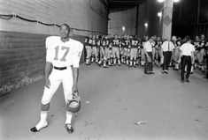 an old black and white photo of a football player holding a helmet in front of a group of people