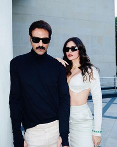 a man and woman standing next to each other in front of a white wall wearing sunglasses