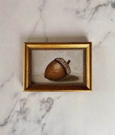 an acorn in a gold frame on a white marble countertop, with a painting hanging above it