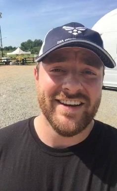 a man with a beard wearing a black shirt and a baseball cap smiles at the camera