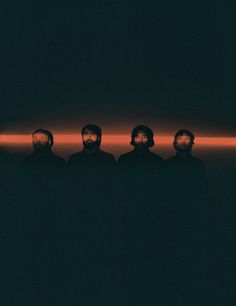 three men with beards standing in front of a black background and red light behind them