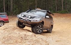 two cars are parked in the dirt near some trees and bushes, one is off - road