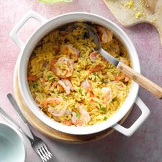 a pot filled with rice and shrimp on top of a pink table cloth next to utensils