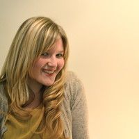 a woman with long blonde hair smiling and holding a cell phone to her ear while standing in front of a white wall