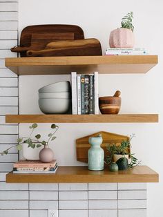 three wooden shelves with books, bowls and other items on them in front of a white brick wall