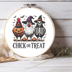 a cross stitch halloween scene with two chickens and a skeleton in a witches hat, on a white wooden background