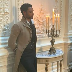 a man leaning against a wall next to a table with candles on it and a chandelier in the background