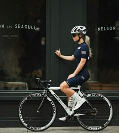 a woman riding a bike down the street in front of a store with her hand up