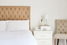 a bed with white sheets and pillows in a bedroom next to a chair, lamp and nightstand