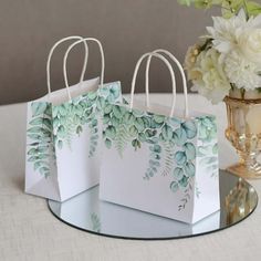 three white bags with green leaves on them sitting next to a vase filled with flowers